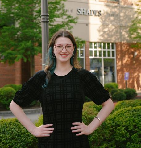 Smiling woman with glasses and blue-streaked hair, hands on hips, in front of a shop.
